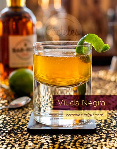 Vaso de cóctel Viuda Negra de color ámbar burbujeante, decorado con una larga espiral de cáscara de lima, descansando sobre un posavasos negro y mantel de estrellas doradas brillantes. Fondo borroso con botella de licor y lima entera.